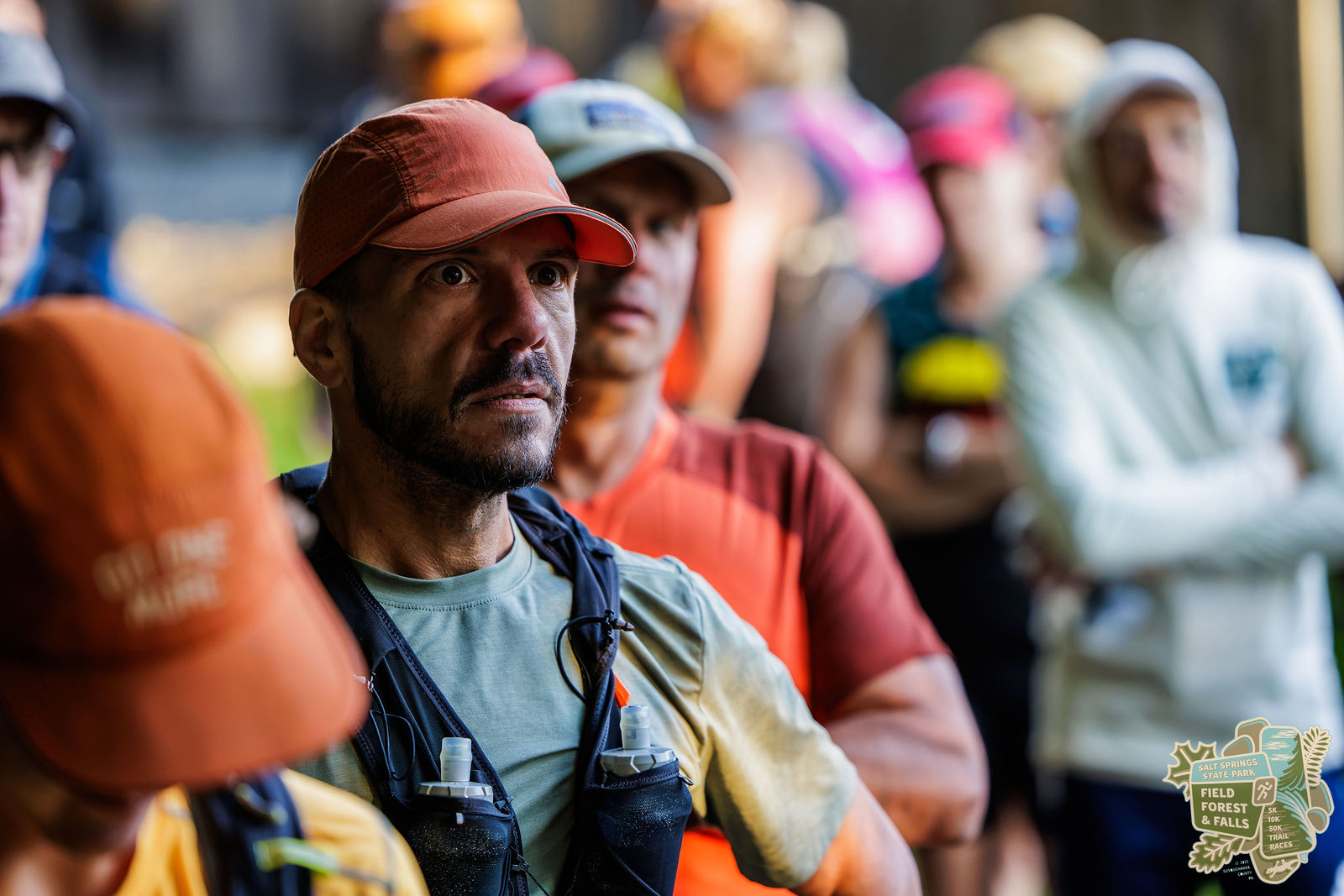 A middle-aged man in an orange trail cap and dark beard listening intently while standing in a crowd.